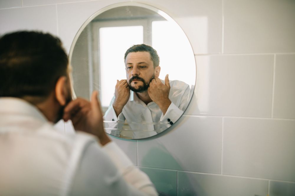Man grooming his beard while looking in a round bathroom mirror, wearing a white shirt.