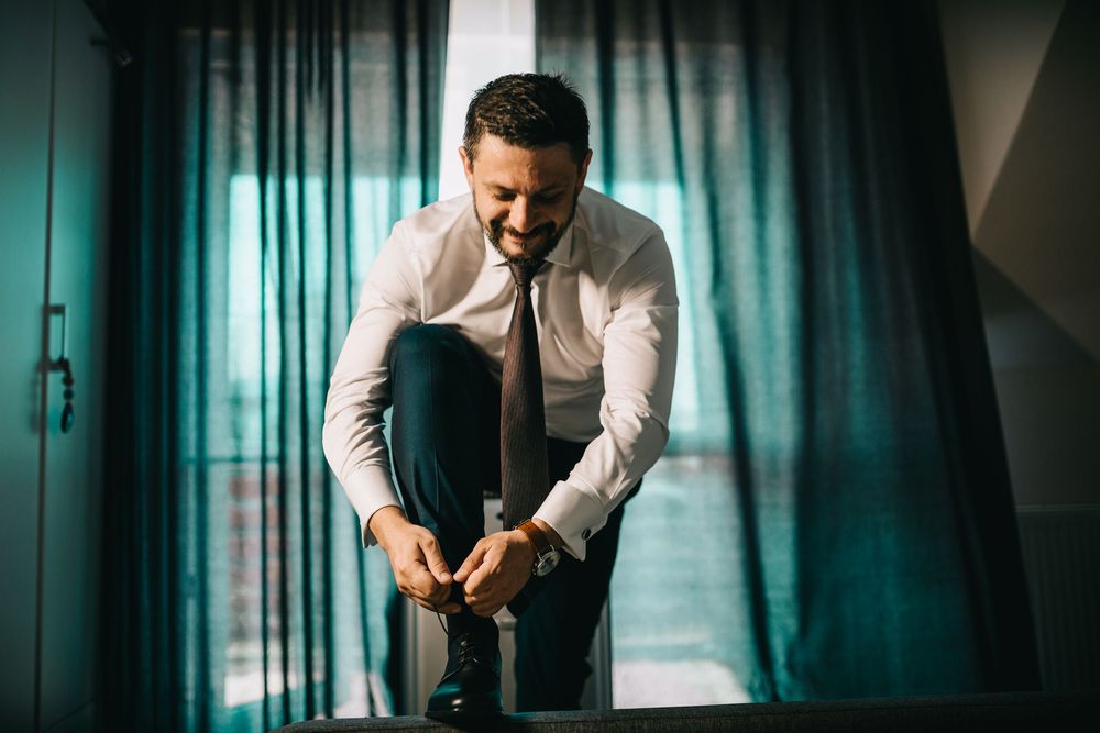 Smiling businessman in white shirt and tie tying his shoes while sitting on a bench near teal curtains.