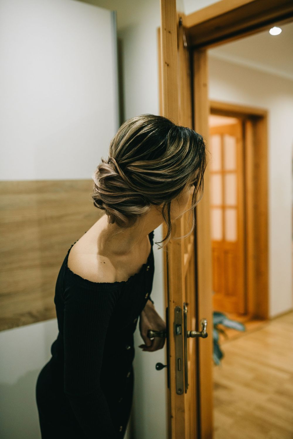 Elegant woman in black off-shoulder dress with updo hairstyle opening a wooden interior door.