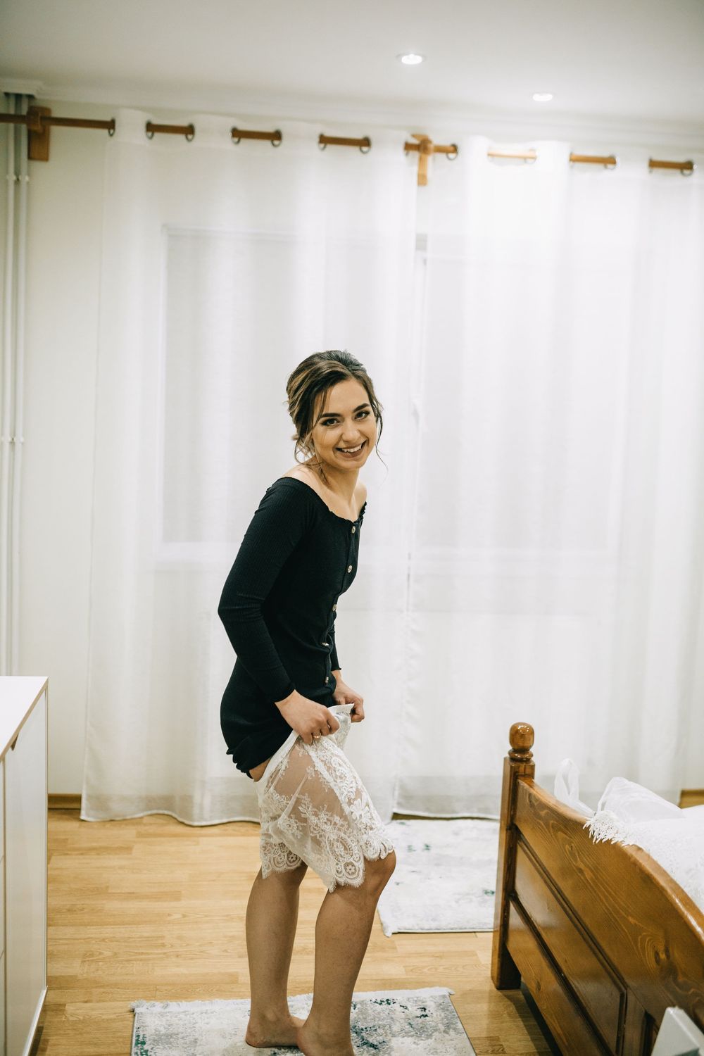 Smiling young woman in black top and white lace skirt standing barefoot in a bright bedroom.