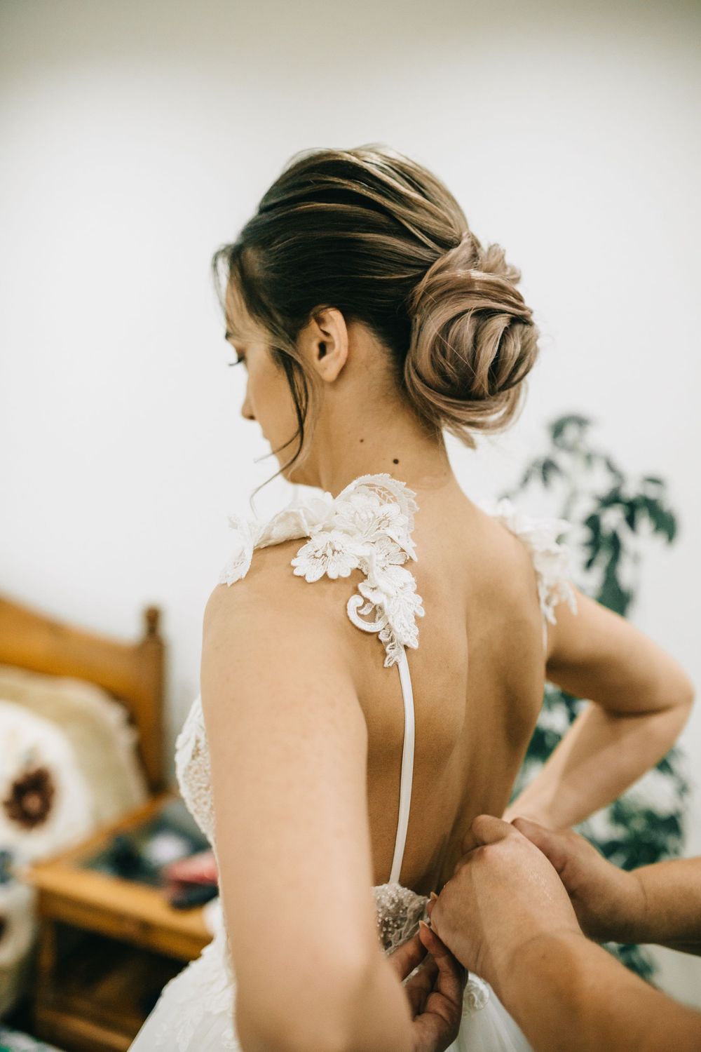 Bride in lace backless wedding gown with elegant updo hairstyle being buttoned up before ceremony.