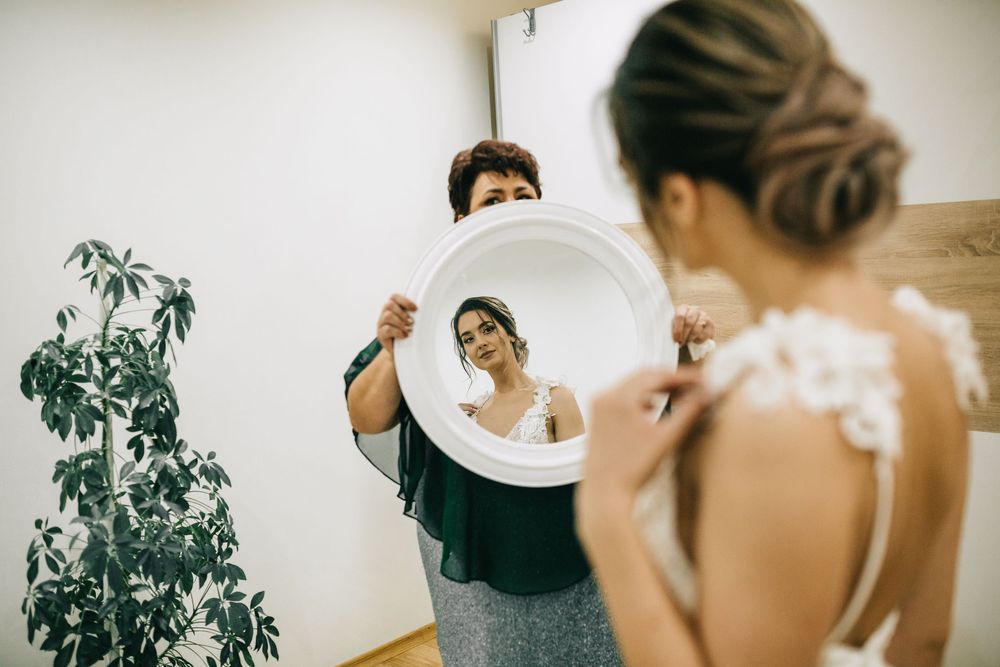 Bride in white gown views her back in a round mirror held by a woman during wedding preparations.