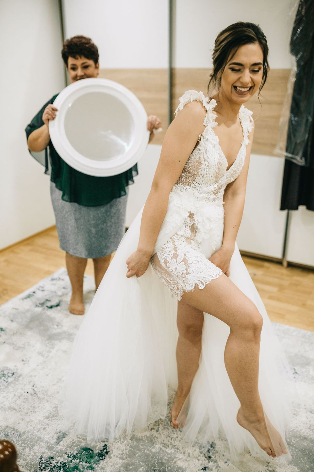 Bride laughing in lace tulle wedding dress while woman holds mirror during bridal preparation.