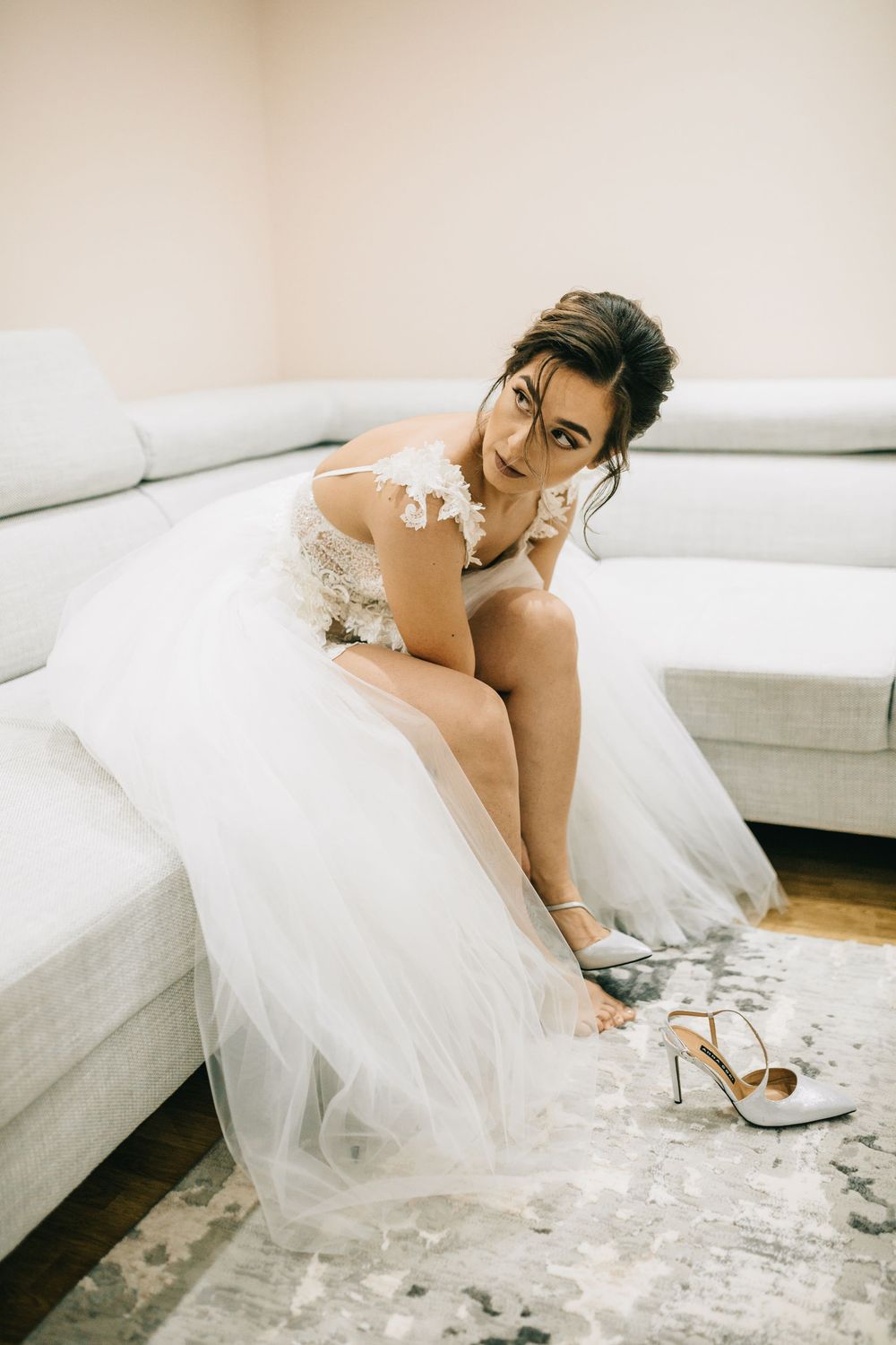 Bride in a white tulle wedding gown putting on her heels while seated on a grey sofa.