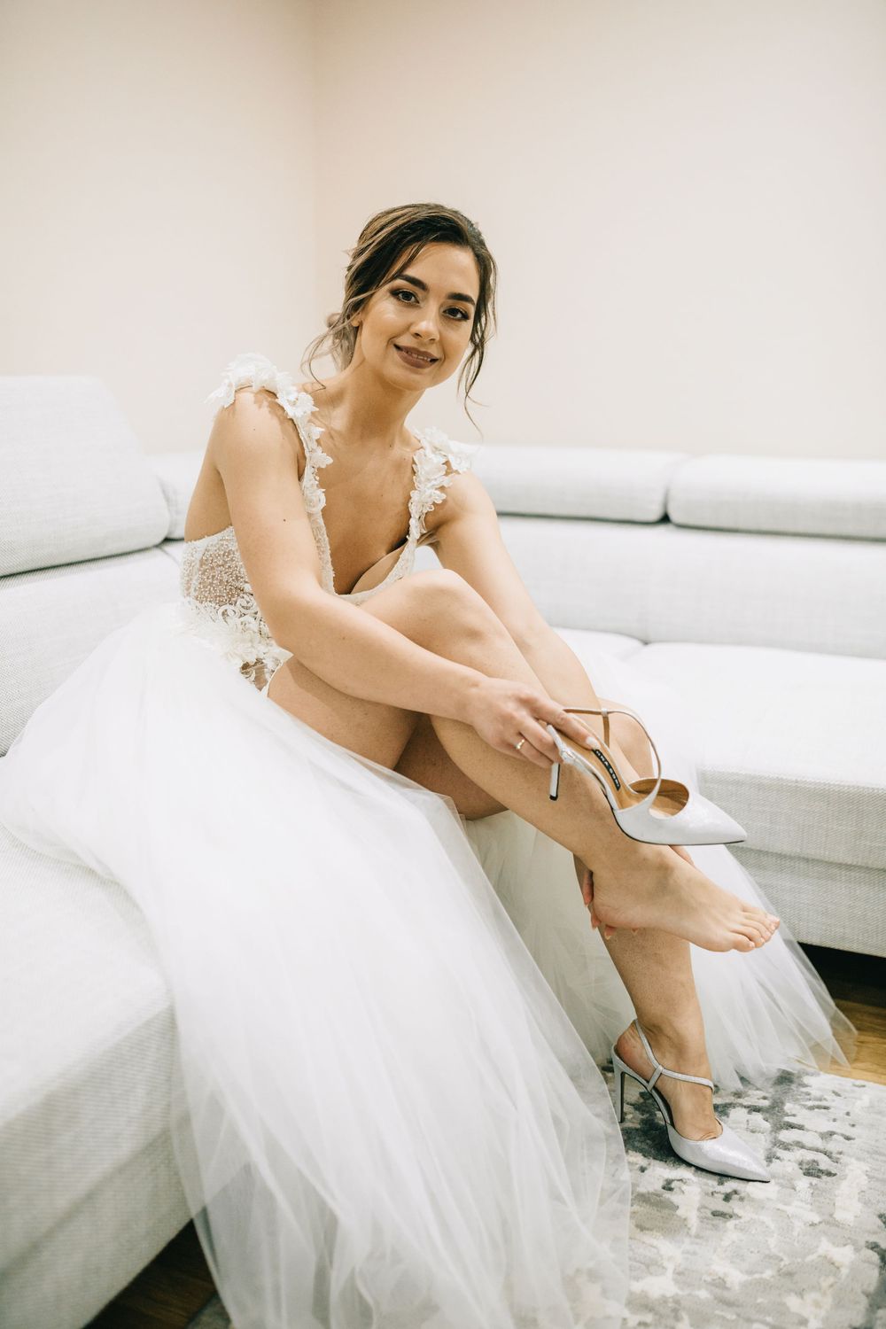 Bride in white tulle wedding dress putting on heeled shoes while sitting on gray sofa.