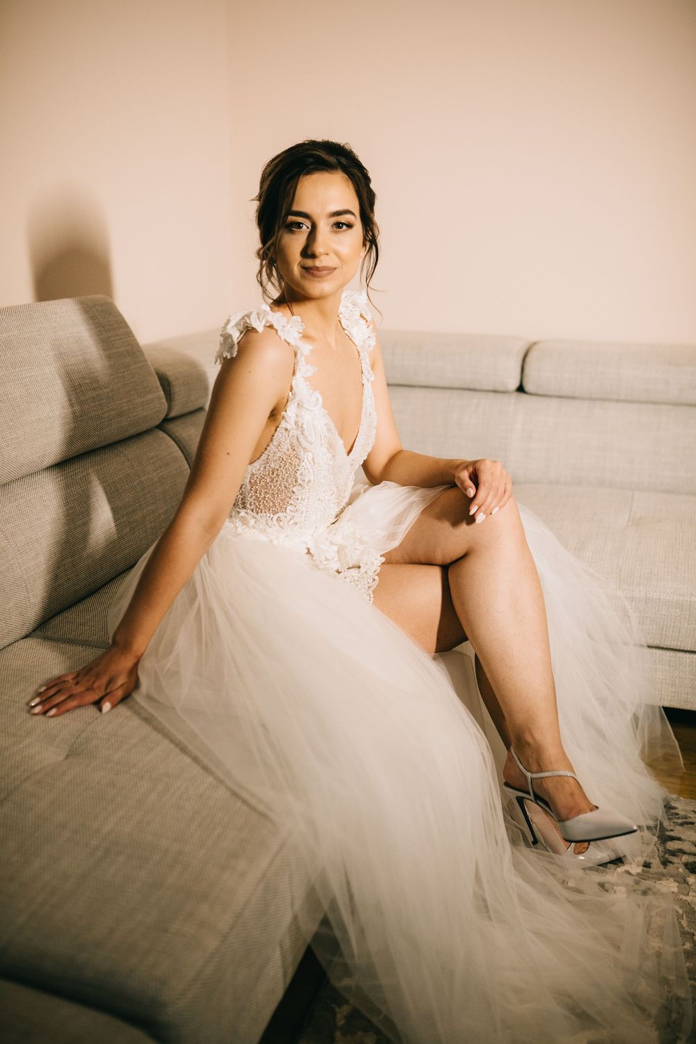 Elegant bride in lace tulle wedding gown sitting on gray sofa wearing silver heels.