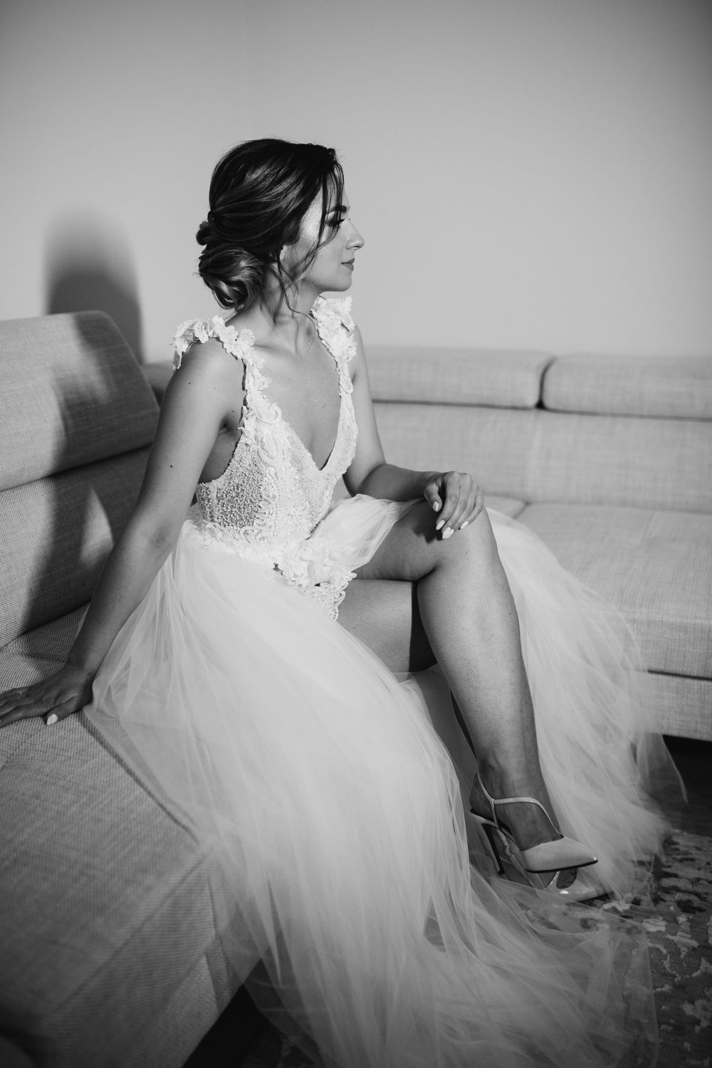 Elegant bride in a lace tulle wedding gown sitting on a sofa in a black and white portrait photo.