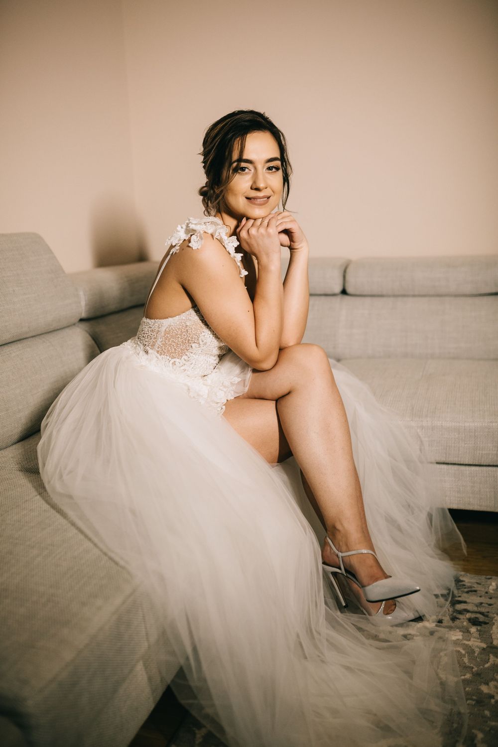 Bride in white lace tulle wedding dress sitting gracefully on gray sofa with hands clasped under chin.