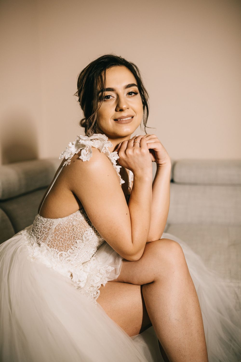 Beautiful bride in a lace and tulle wedding dress smiling softly while seated on a grey sofa.