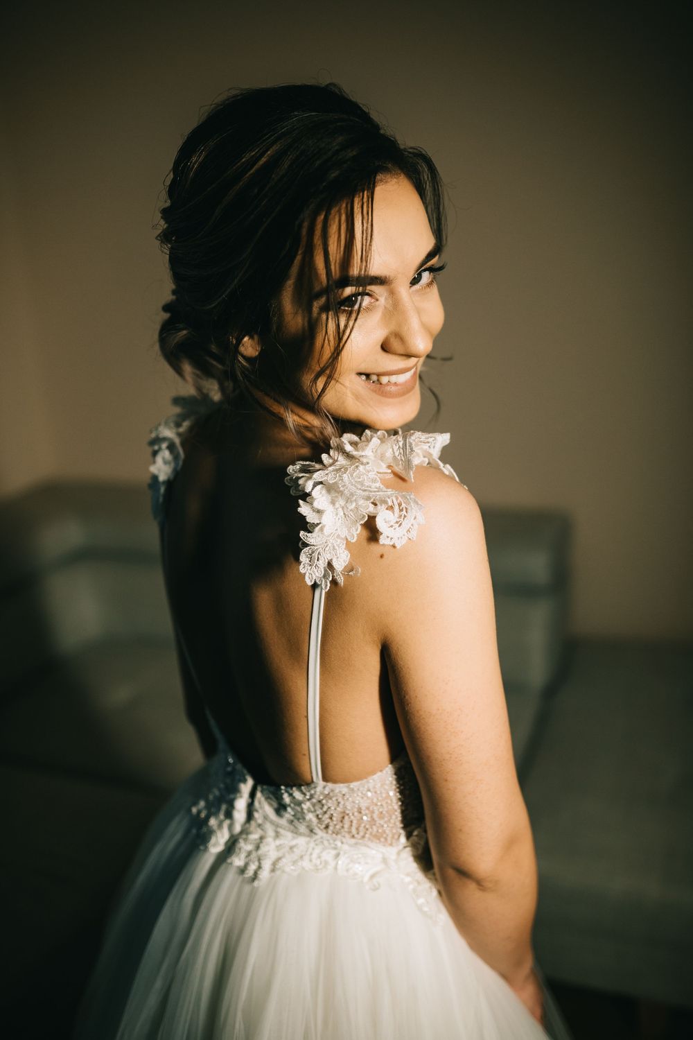 Smiling bride in backless lace wedding gown with floral shoulder detail poses elegantly indoors.