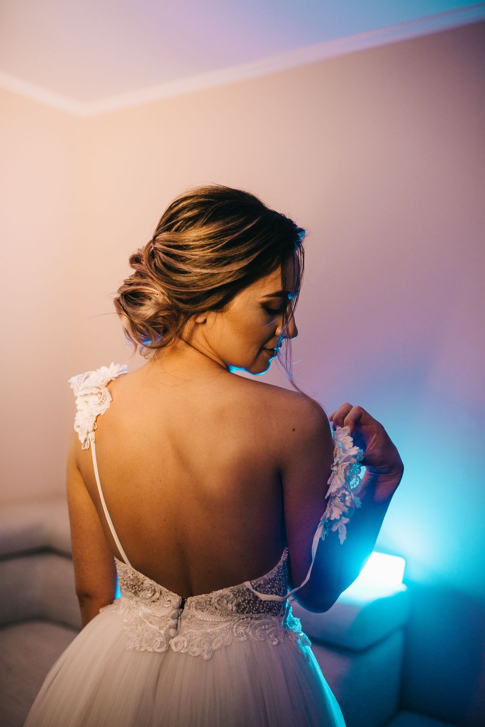 Elegant bride in a backless lace wedding gown adjusting her dress strap in a softly lit room.
