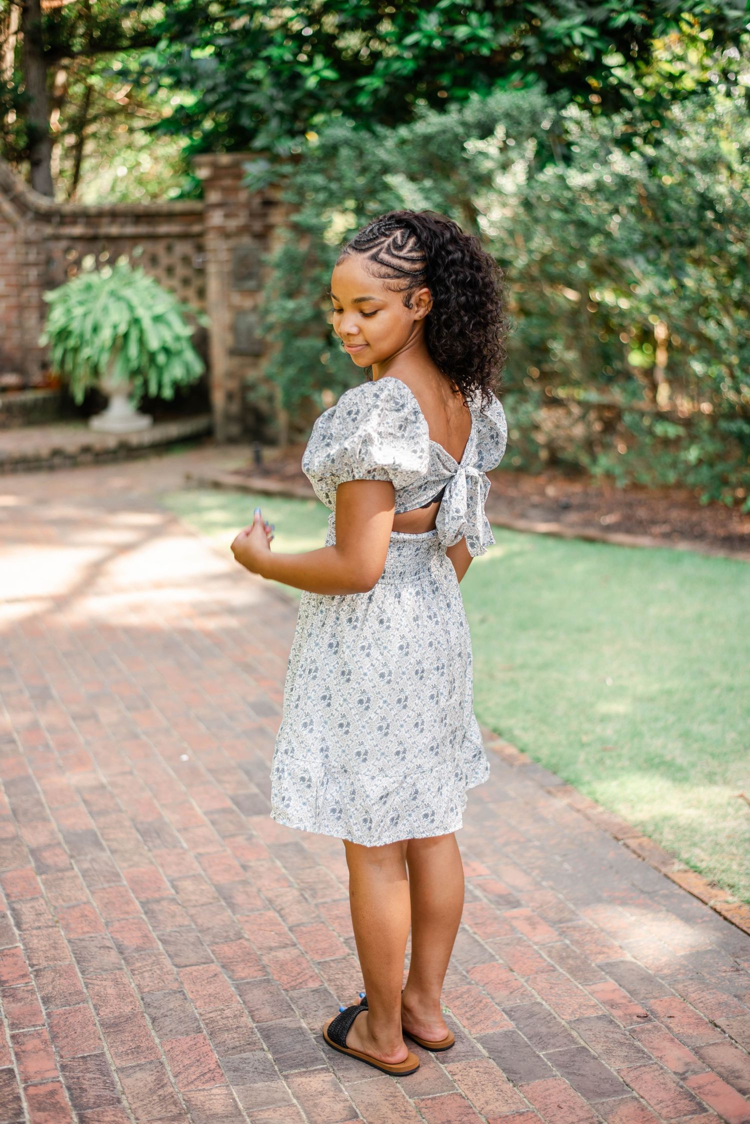 Young girl in white floral dress with bow back, standing on brick path in lush garden setting.