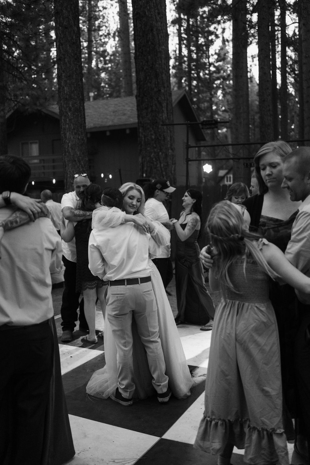 Black and white photo of couples dancing outdoors at a wedding reception surrounded by tall pine trees.