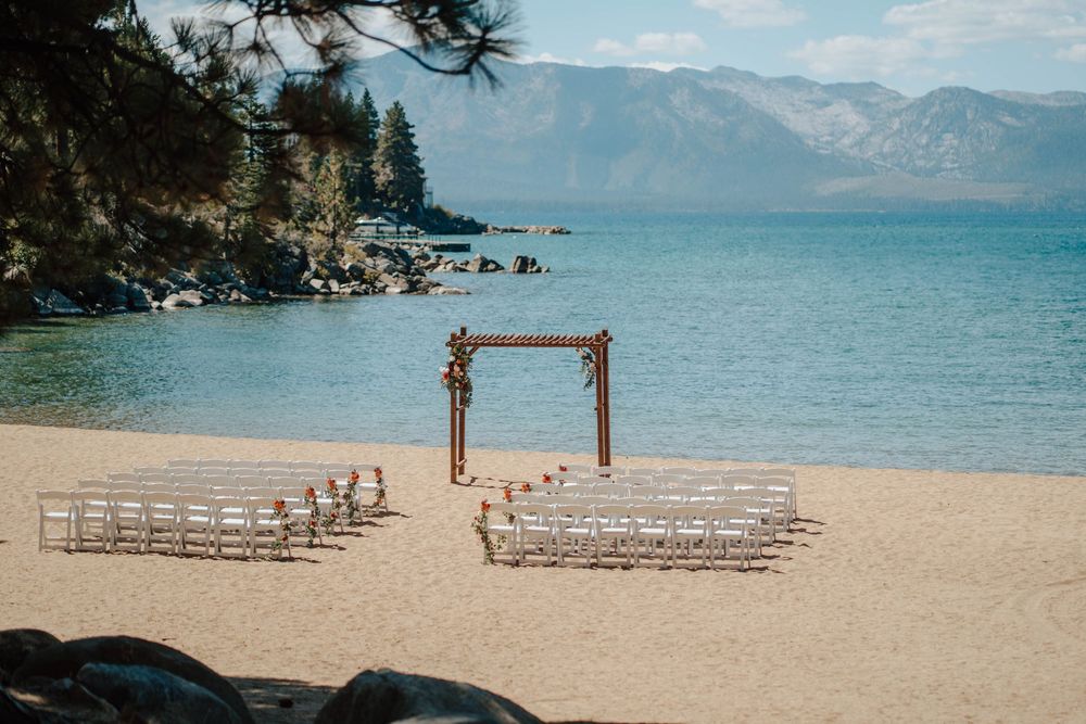 Elegant outdoor beach wedding setup with white chairs and wooden arch overlooking Lake Tahoe mountains.