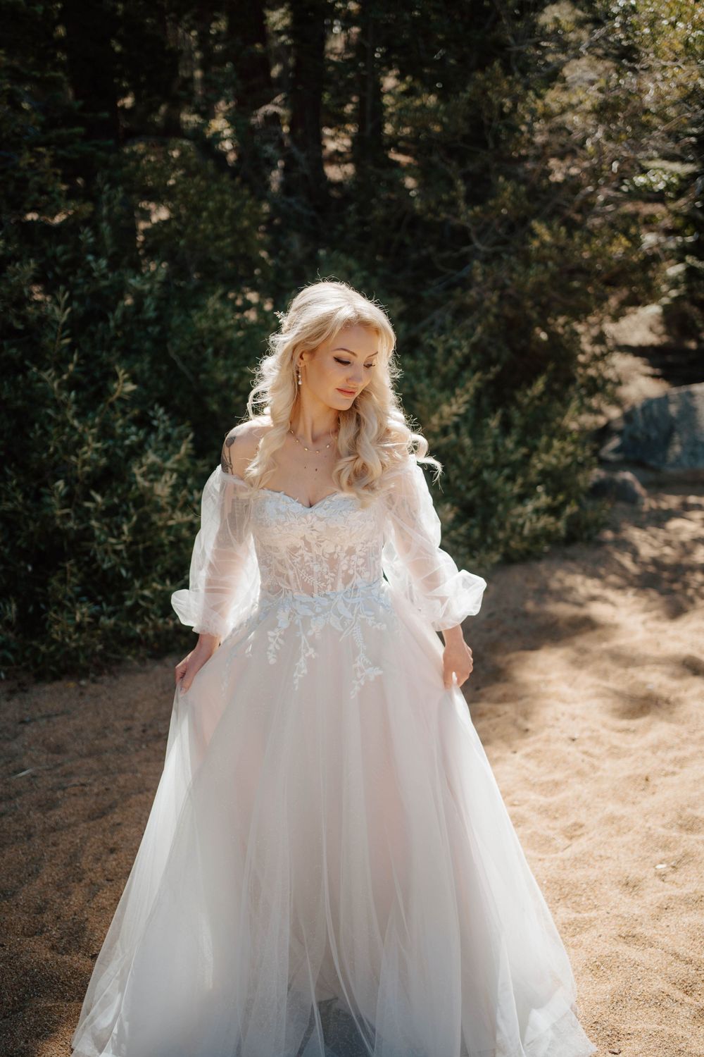 Blonde bride in a flowing white lace tulle ball gown poses outdoors among green trees on sandy ground.