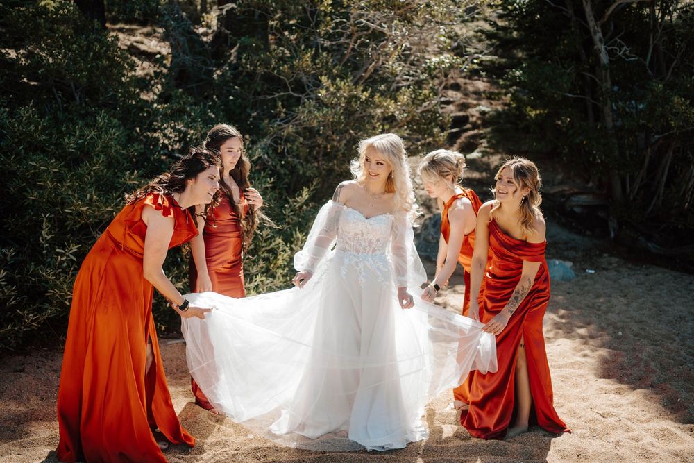 Bride in white gown with bridesmaids in rust orange dresses helping arrange her train outdoors in natural setting.