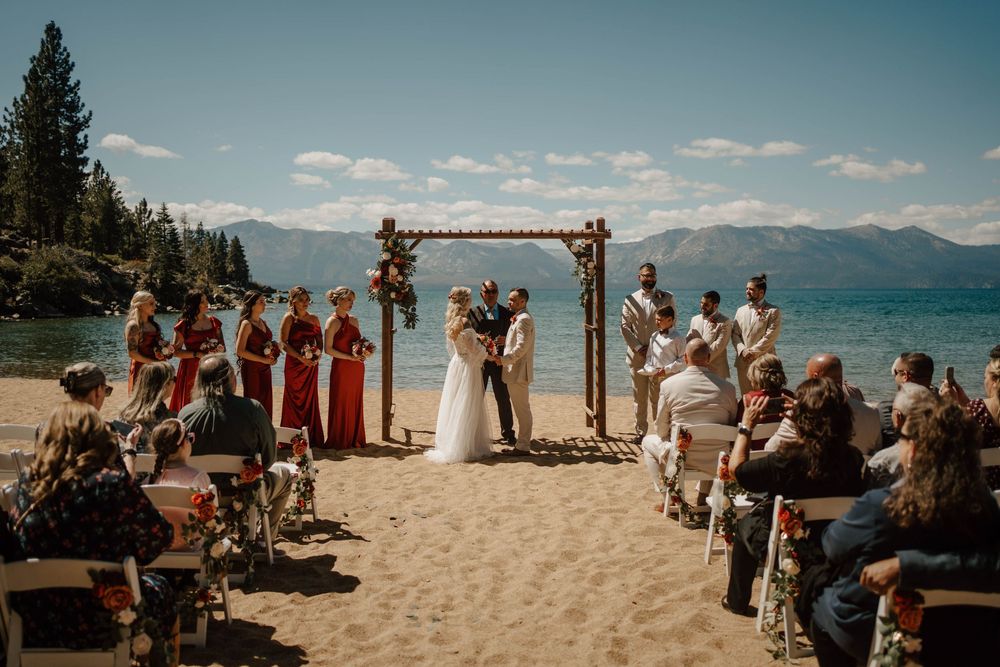 Beach wedding ceremony at Lake Tahoe with couple at rustic arch, bridal party in red, guests seated on sandy shore with mountain views.