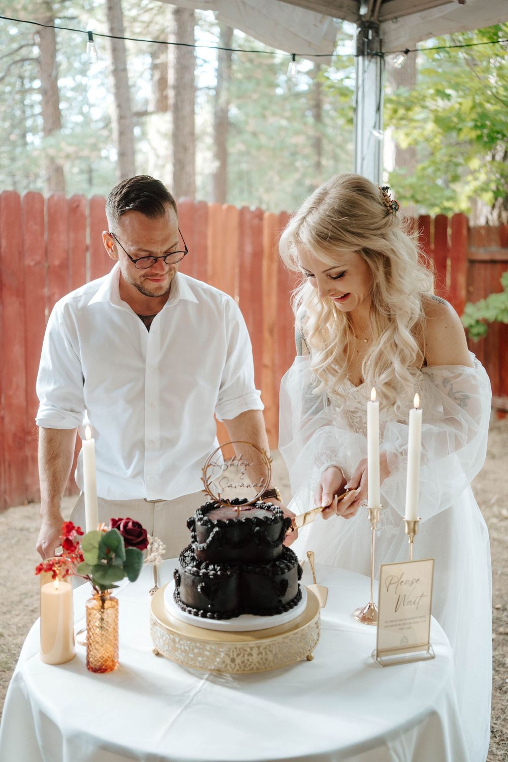 Bride and groom cutting a dark two-tier wedding cake at an outdoor reception table with candles and floral arrangements.