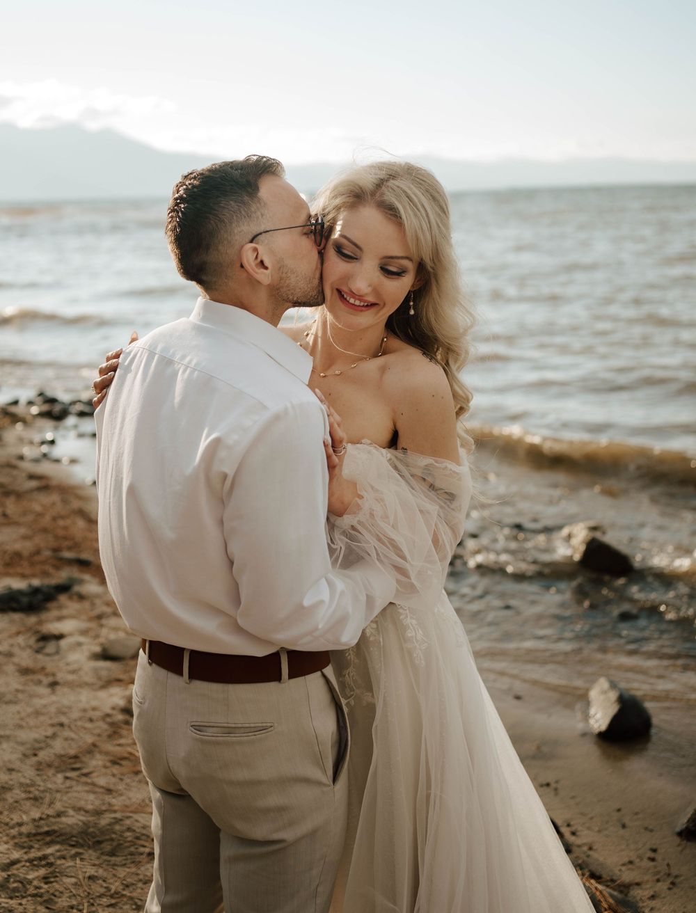 A couple shares a romantic moment on a sandy beach shoreline, with the groom kissing the bride in her elegant off-shoulder gown.