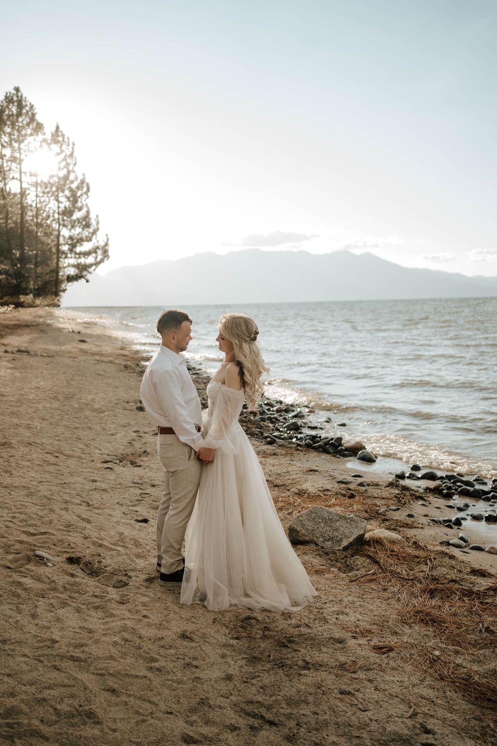 Bride and groom holding hands on a sandy lakeside beach at sunset with mountains in the background.