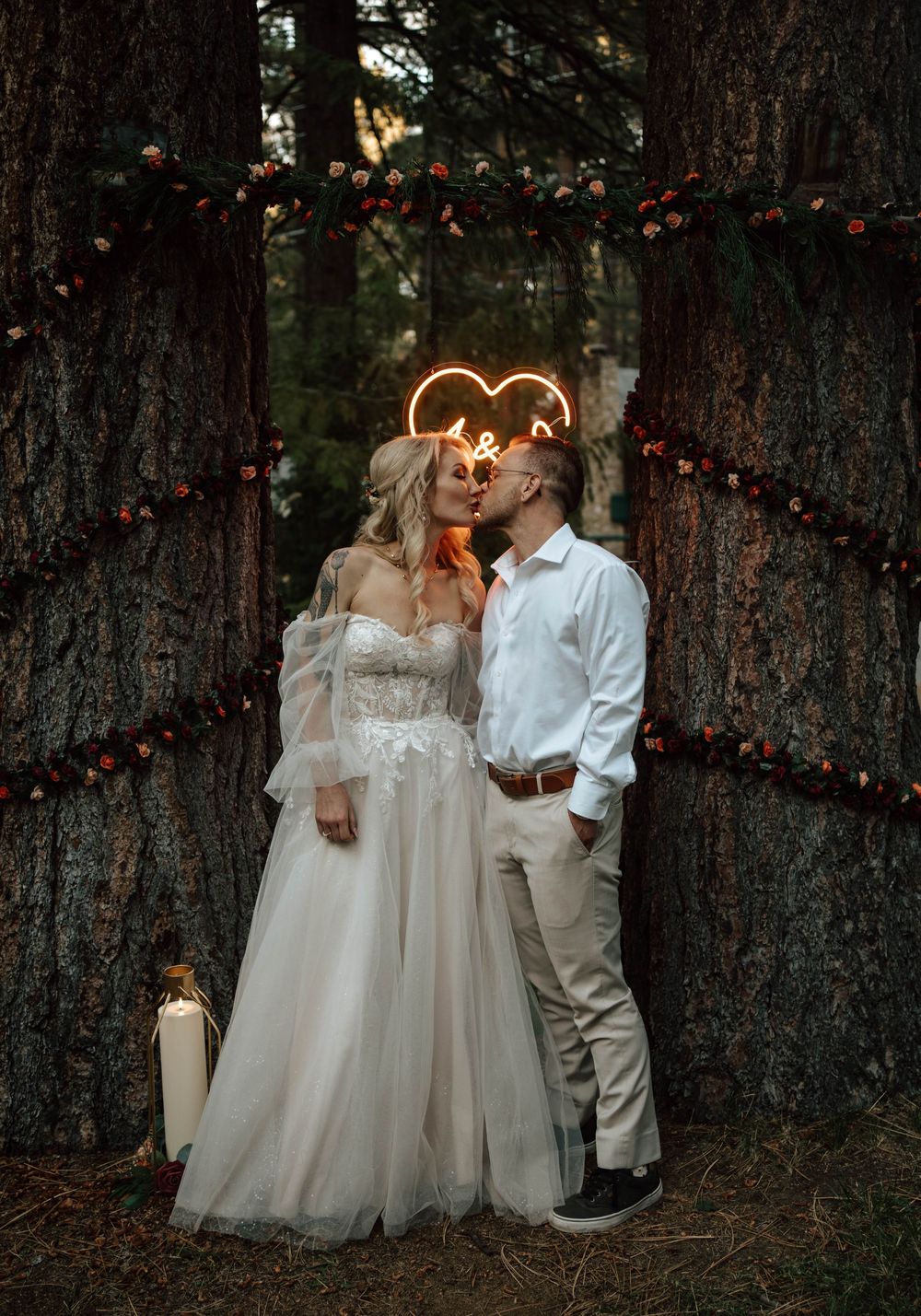 A bride and groom share a romantic kiss in a forest wedding setting with neon heart sign and floral garlands on trees.