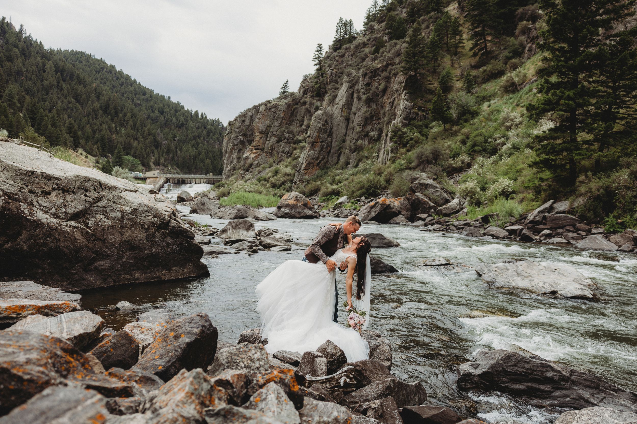 Couple sharing a romantic kiss in wedding attire on rocky riverbank with rushing water and pine-covered cliffs in background.
