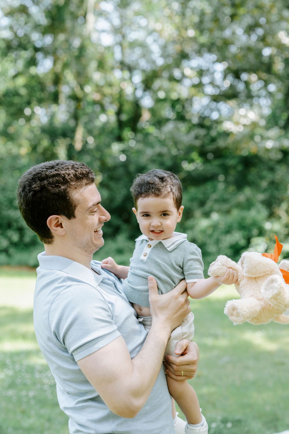 A smiling father holds his happy toddler and a stuffed bear in a sunny green outdoor setting.