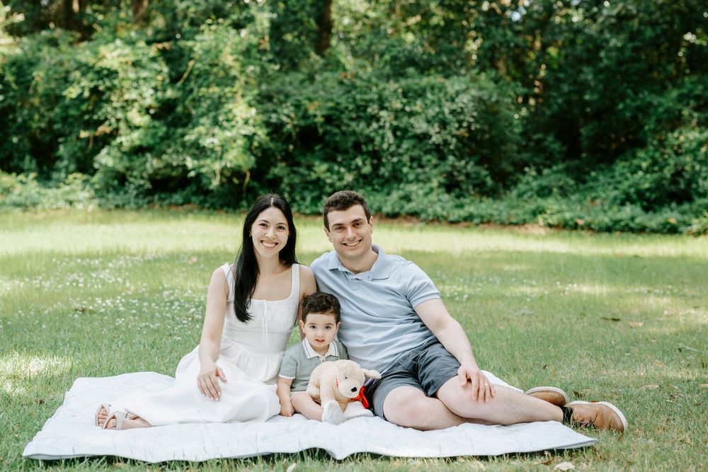 Family of three sitting on white blanket in green park, parents smiling with toddler between them.