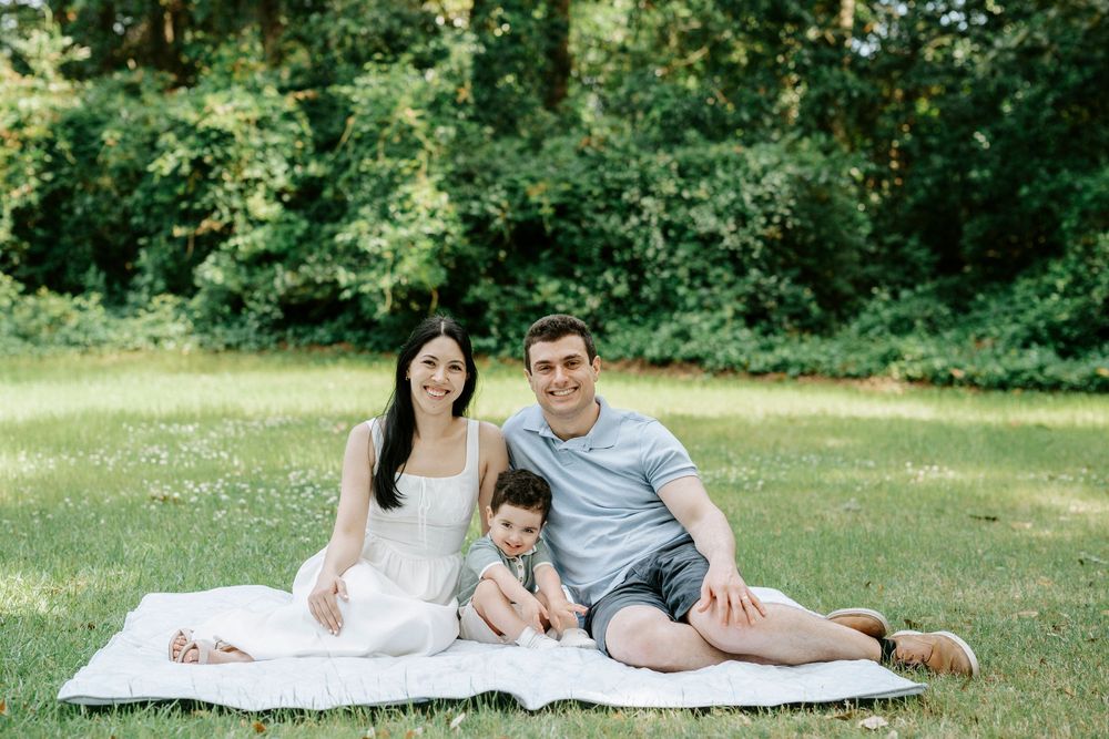 A happy family of three sitting on a white blanket in a lush green park, smiling for a outdoor portrait photo.