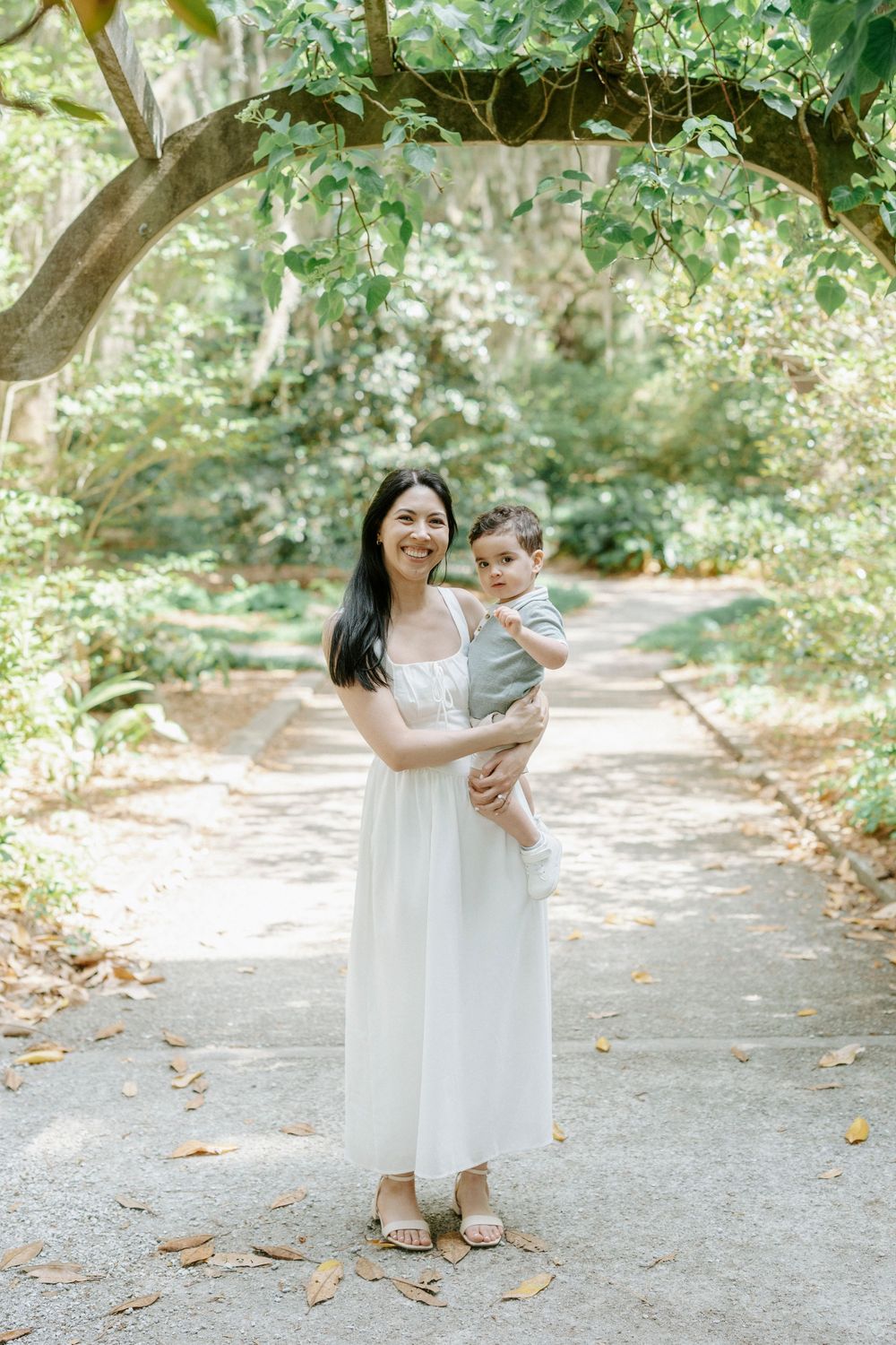 A smiling woman in a white dress holds a baby in a lush green park pathway.