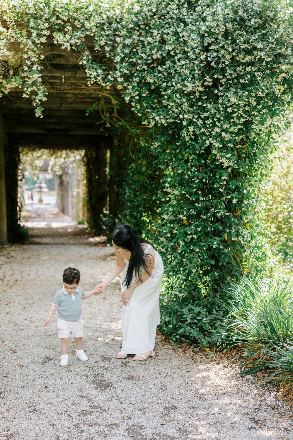 A mother in a white dress holds her toddler son's hand along a lush green garden pergola pathway.