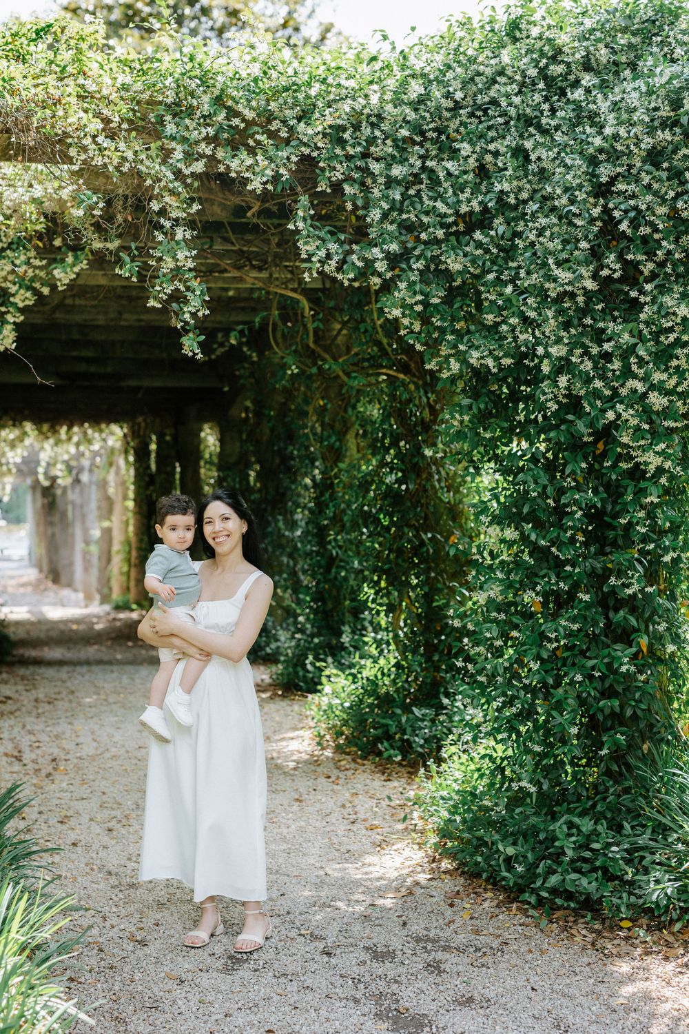 A smiling woman in a white dress holds a baby near a lush ivy-covered pergola in a sunlit garden.
