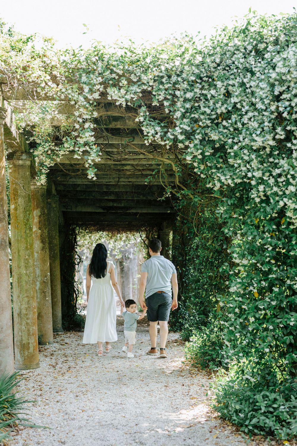 Family of three walking hand-in-hand through ivy-covered garden archway on sunny day.
