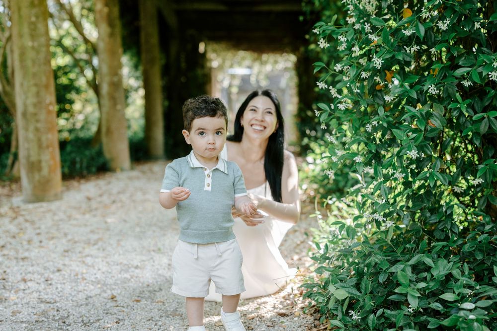 A smiling woman crouches beside a young boy on a garden path surrounded by lush greenery and trees.