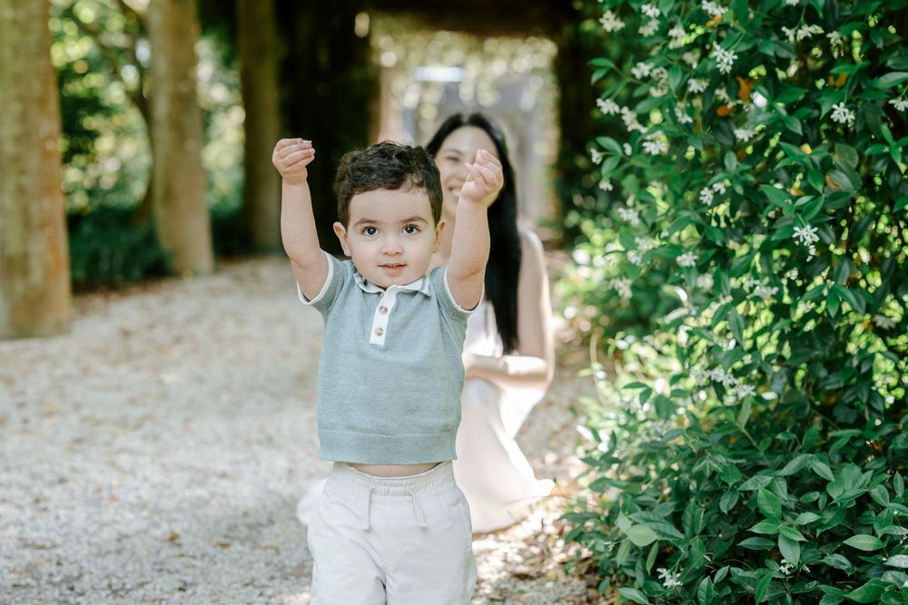 Toddler boy walking with arms raised on a garden path, with a woman smiling in the background.