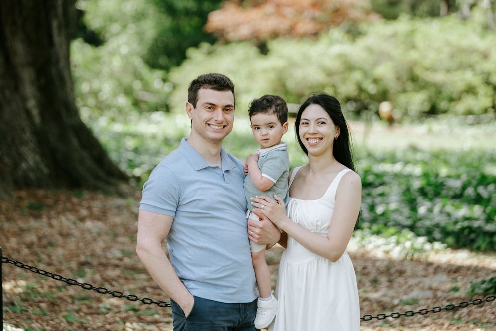Smiling family of three posing outdoors in lush green park, parents holding young toddler.