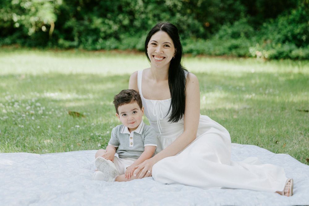 Young mother in white dress sitting on blanket in green park with her smiling toddler son.