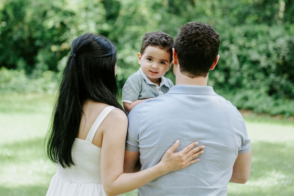 Family of three with mother in white dress and father holding toddler son in lush green park.