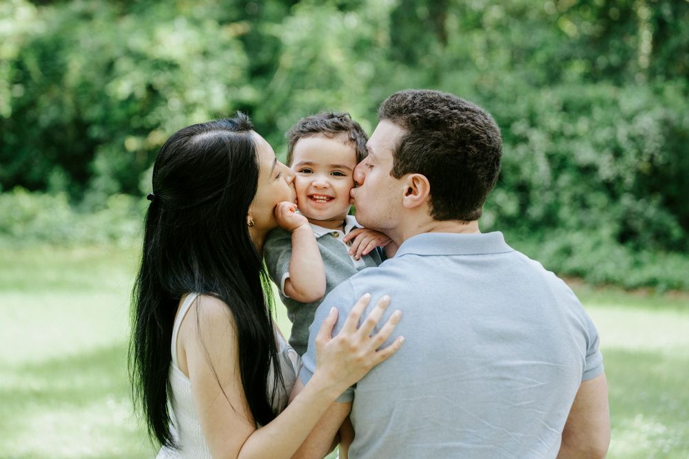 Two parents lovingly kiss their smiling toddler on the cheeks in a lush green outdoor setting.