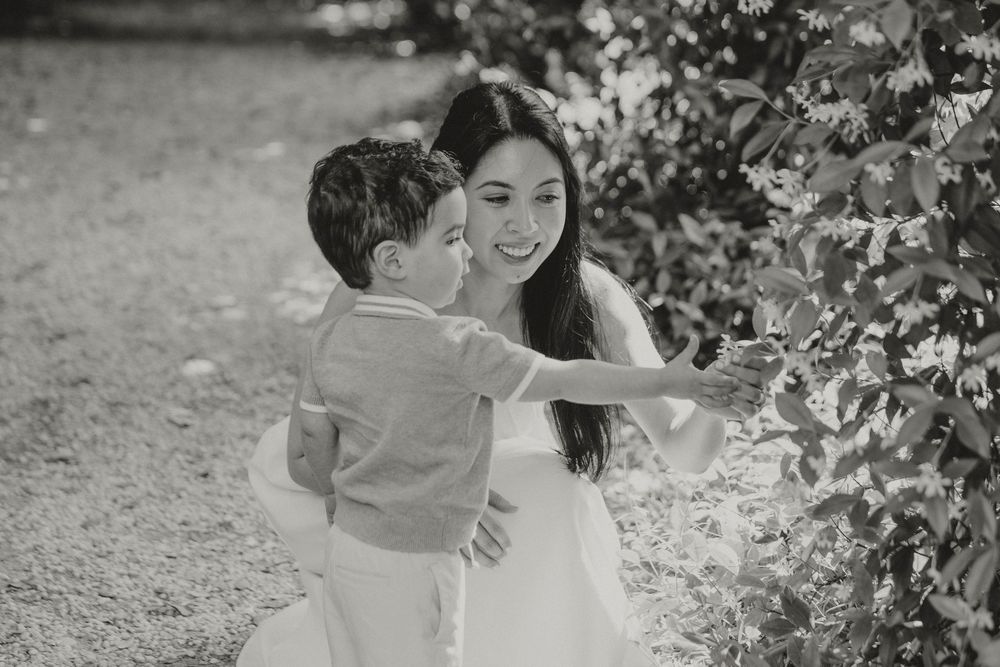 A smiling mother crouches to embrace her young son near green foliage in a black and white photo.
