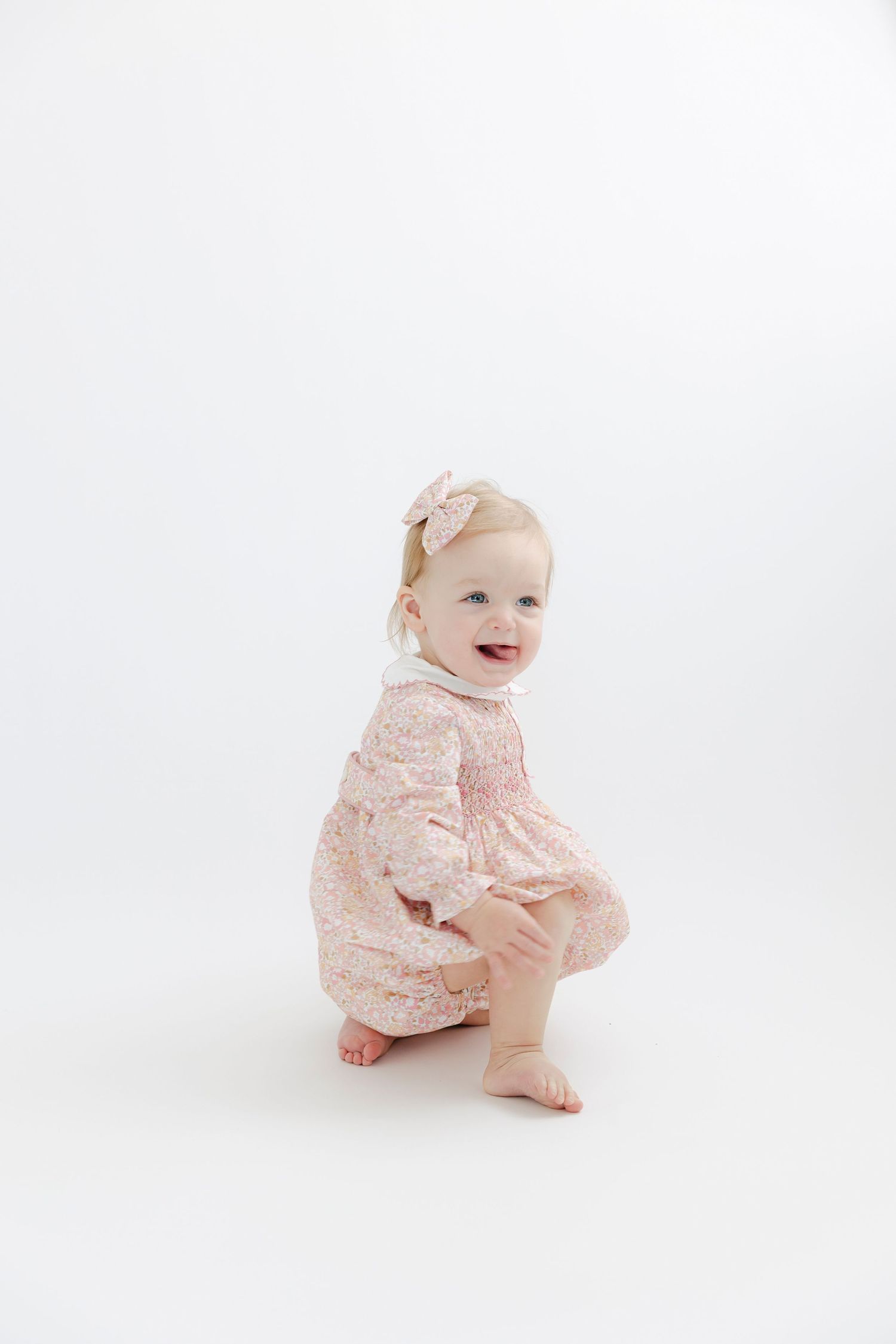 Smiling toddler in pink floral romper crouching on white background with hair bow accessory.