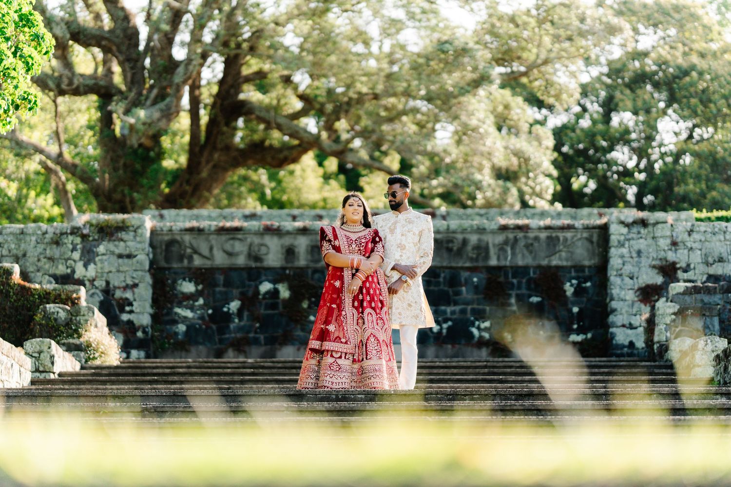 Indian bride in red lehenga and groom in white sherwani pose on stone steps surrounded by lush greenery.