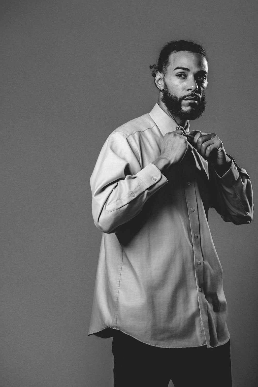 Black and white portrait of a bearded man in a button-up shirt adjusting his collar against a gray background.