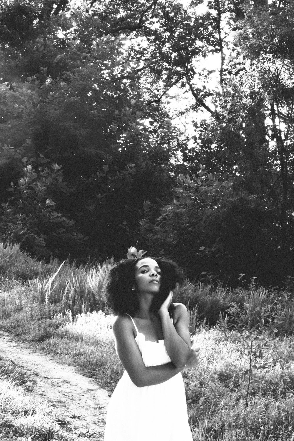 Black and white portrait of a young woman with natural afro hair in a white dress, posing outdoors in a lush forest setting.