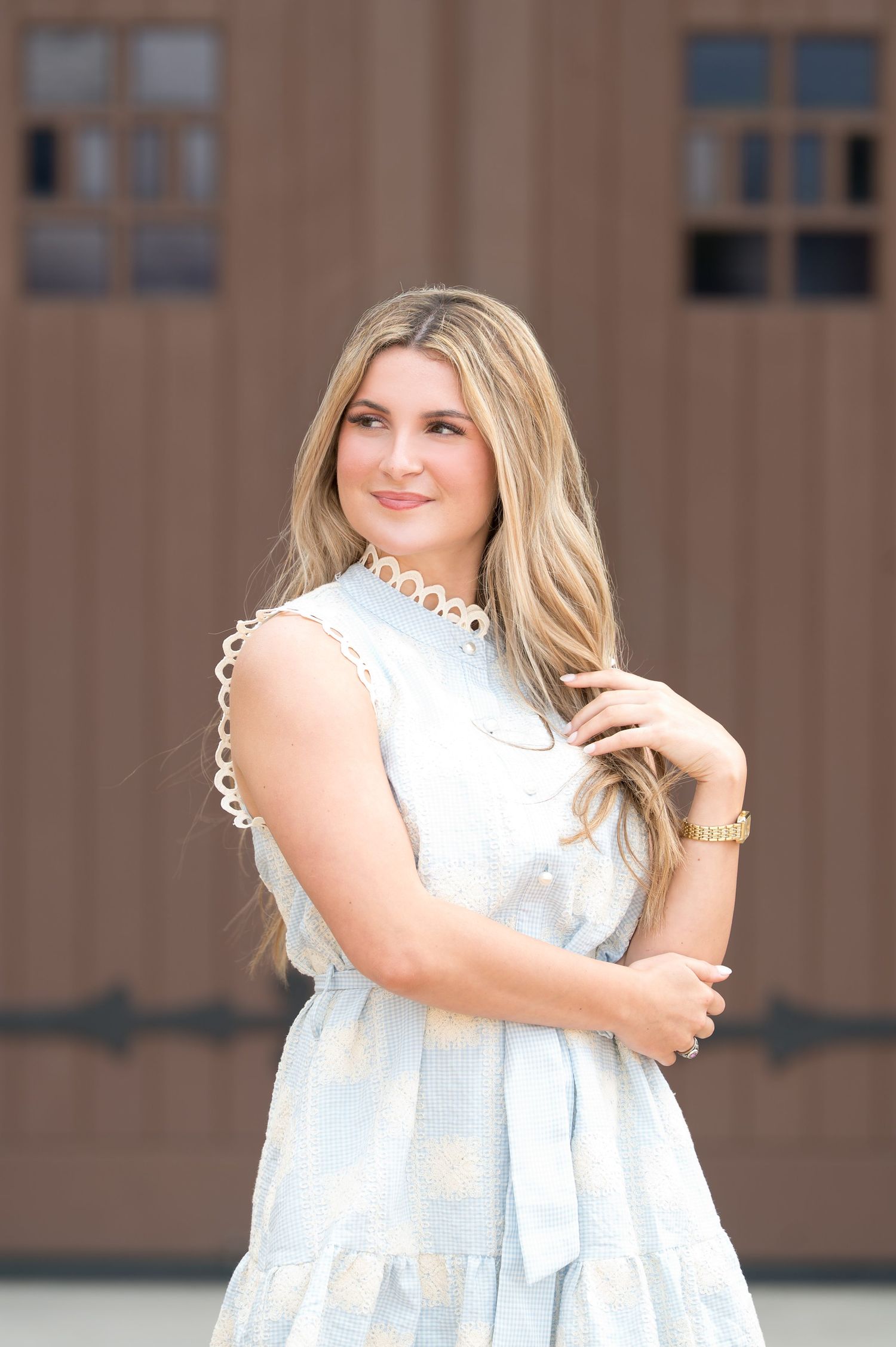 Young woman in light blue sleeveless dress with scalloped trim poses outdoors near wooden barn doors.
