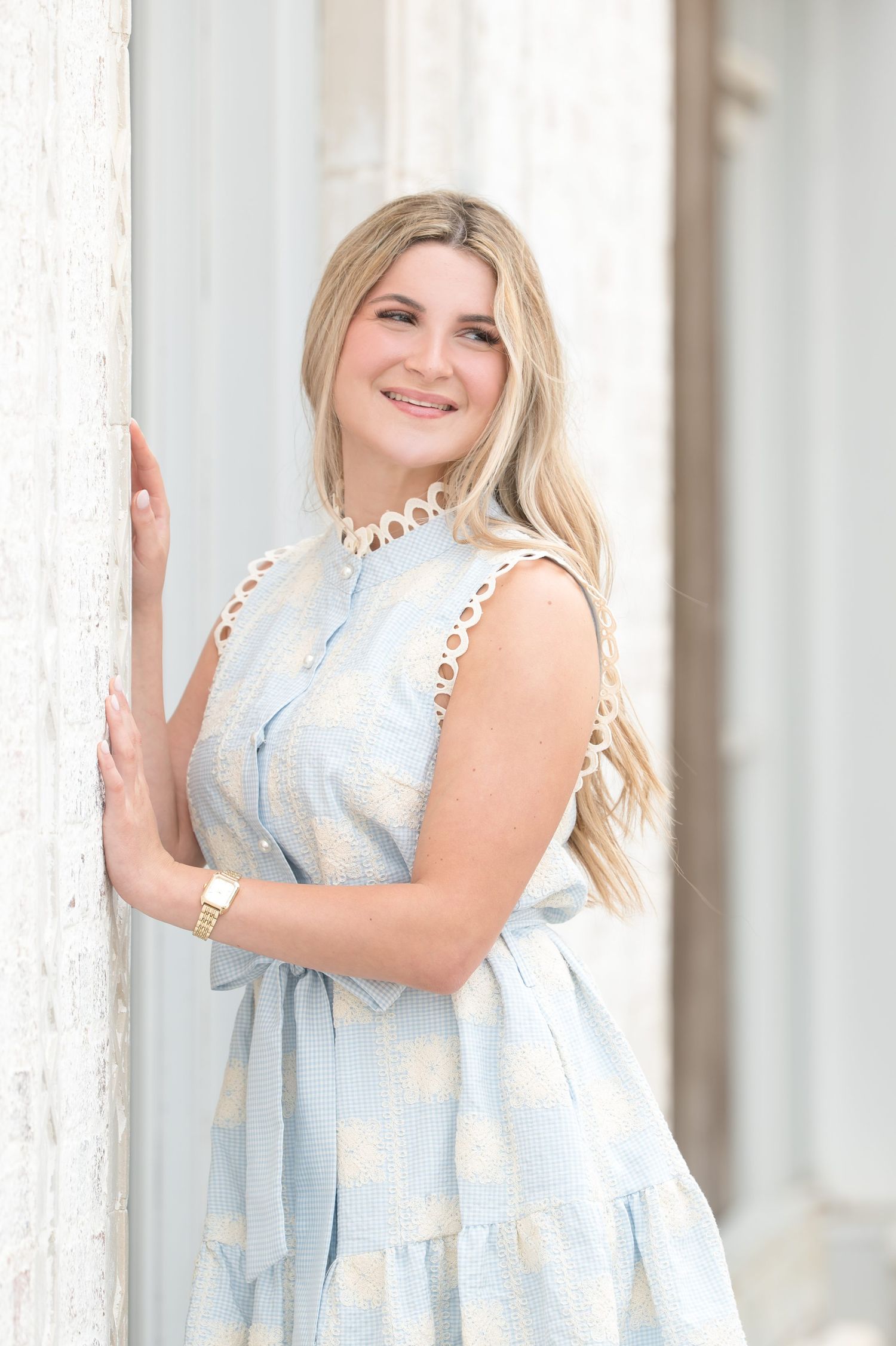 Smiling blonde woman in light blue floral dress leaning against white column in elegant outdoor portrait.