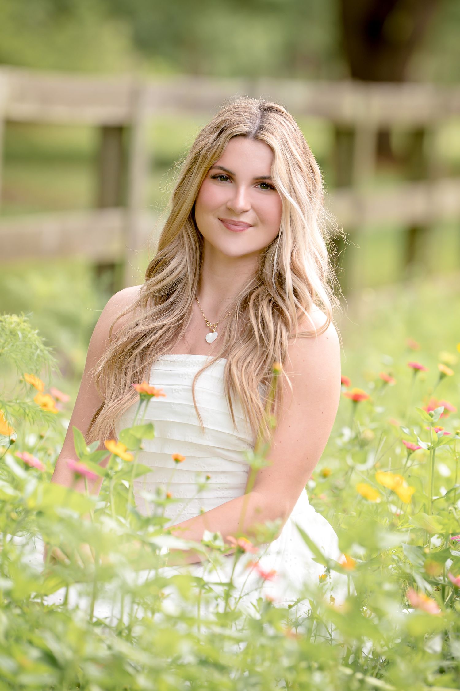 Young blonde woman in white dress sitting among colorful wildflowers in a sunny outdoor garden setting.