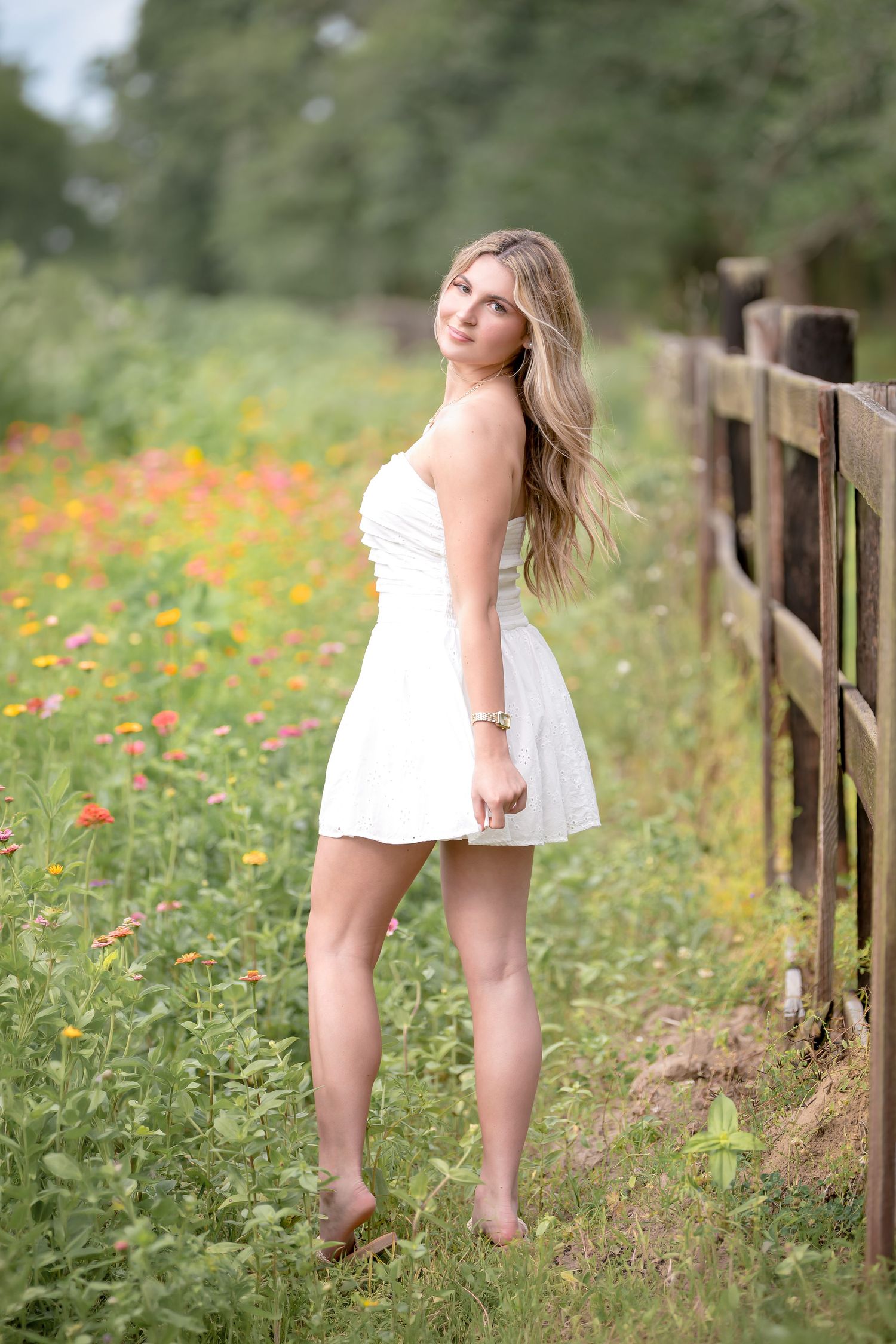 Young woman in white sundress standing by rustic fence in colorful wildflower meadow, long blonde hair flowing.