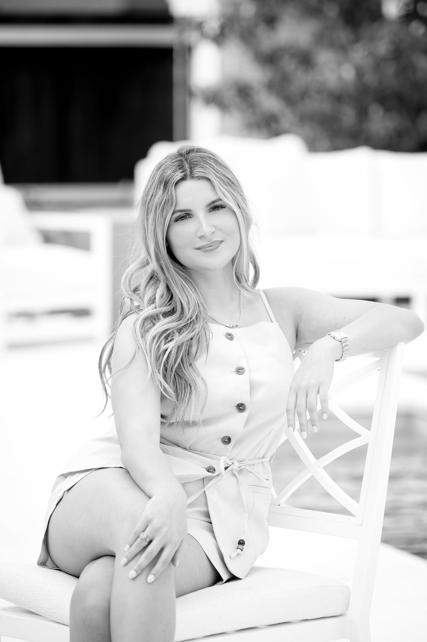 Young blonde woman in button-front dress sits relaxed on white chair in black and white portrait photo.