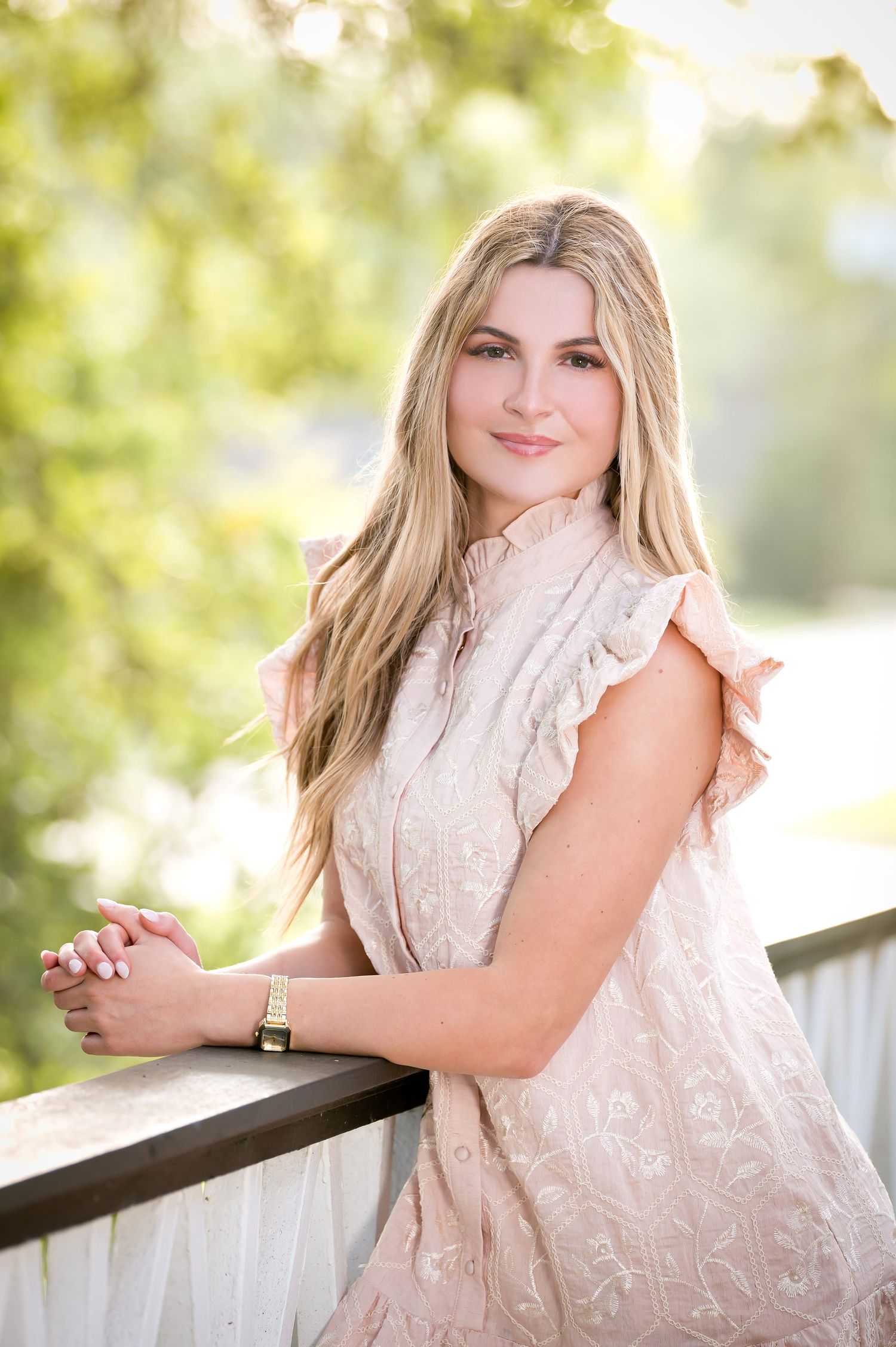 Young woman in blush pink ruffled dress leans on wooden railing surrounded by lush green trees.